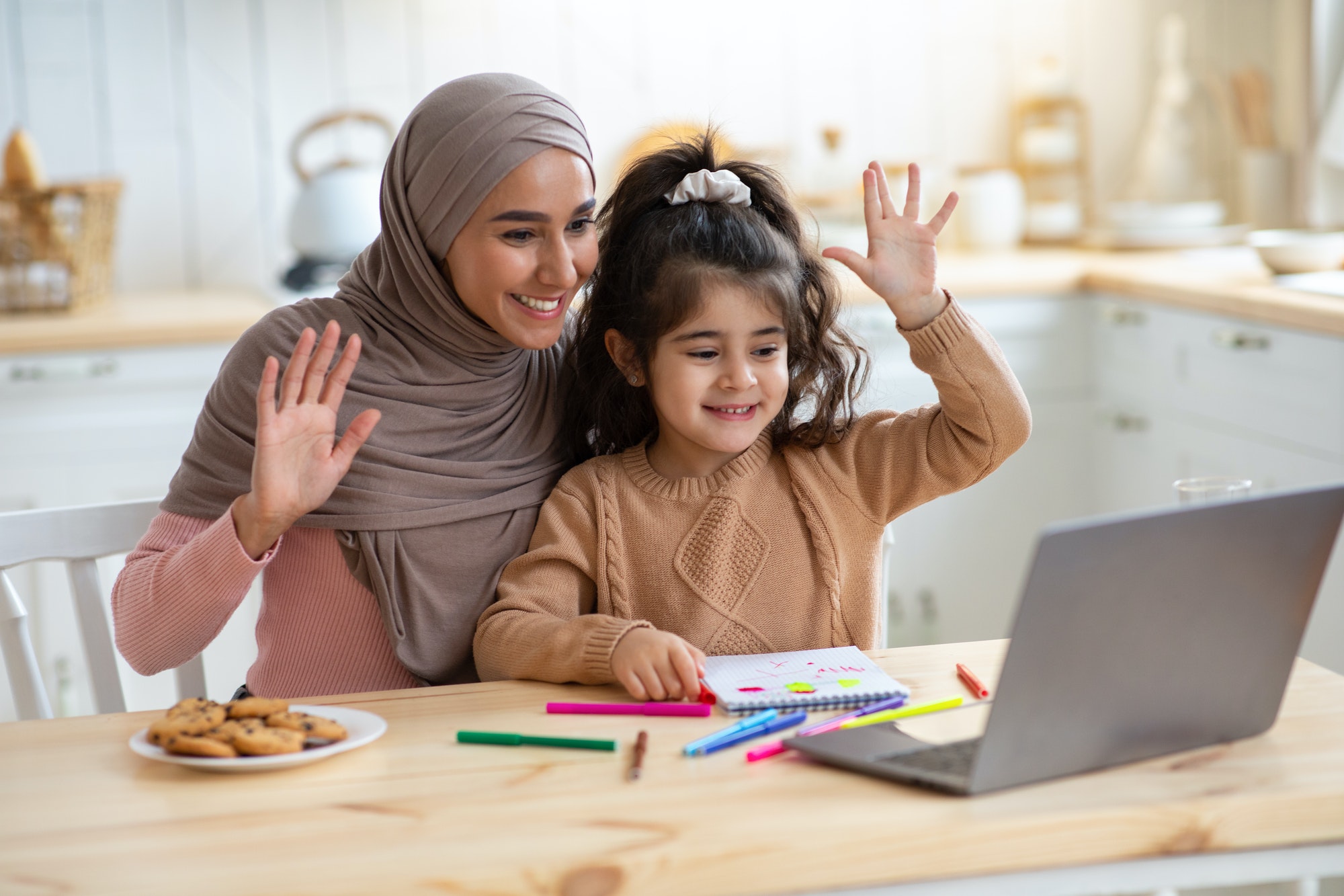 Muslim mother and little daughter making video call on laptop in kitchen